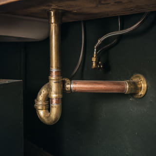 Brass and copper trap pipework under a counter against a dark wall.